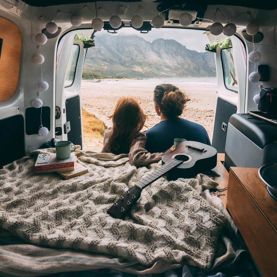 Couple seen from behind on a camper van overlooking mountain scenery at sunrise